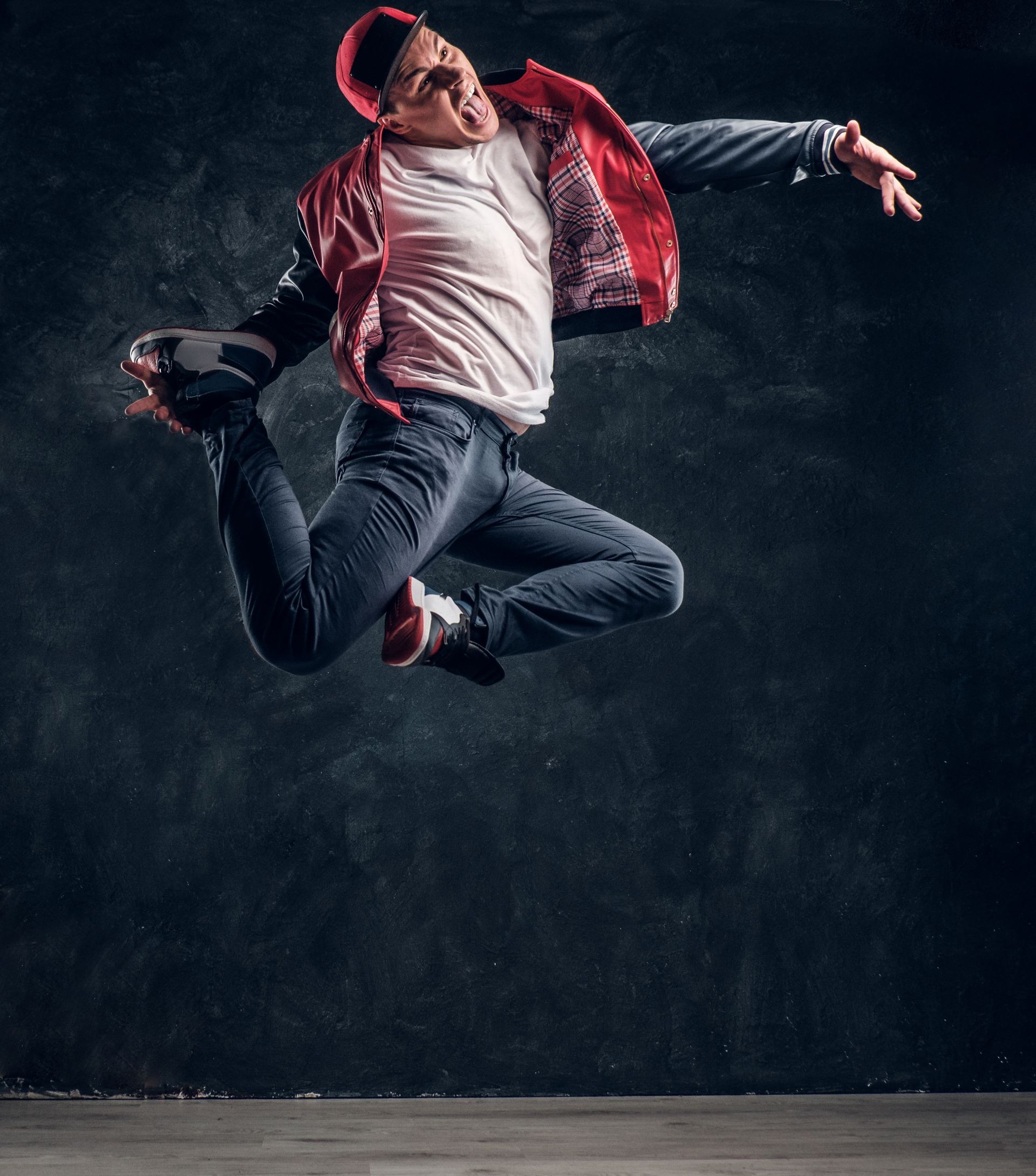 Emotional stylish dressed guy performing break dance jumping. Studio photo against a dark textured wall